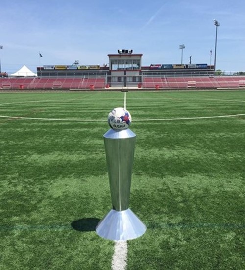 Metal game ball stand on a field with bleachers in the background